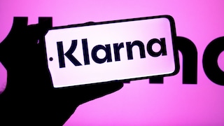 A person holds a smartphone displaying the Klarna logo against a pink-purple background, which also features the Klarna logo. The screen shows the word "Klarna" in black text on a light background.