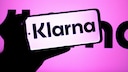 A person holds a smartphone displaying the Klarna logo against a pink-purple background, which also features the Klarna logo. The screen shows the word "Klarna" in black text on a light background.