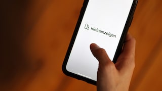A hand holds a smartphone displaying the green logo of Kleinanzeigen. The background features shades of orange and brown.