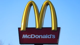 The image shows a large McDonald's sign against a cloudless blue sky. At the top, the yellow, arch-shaped elements of the logo are visible. Below that is a dark red rectangular panel with the white lettering "McDonald's." At the bottom of the image, part of the black support structure of the sign is visible, and to the left, the tips of a bare tree can be seen.