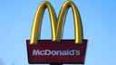 The image shows a large McDonald's sign against a cloudless blue sky. At the top, the yellow, arch-shaped elements of the logo are visible. Below that is a dark red rectangular panel with the white lettering "McDonald's." At the bottom of the image, part of the black support structure of the sign is visible, and to the left, the tips of a bare tree can be seen.
