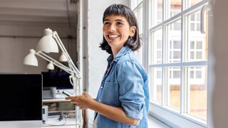 A person in a light denim shirt stands by a window in an office, holding a smartphone. Several monitors, desk lamps, and a notebook are on a desk. The room is bright, illuminated by large windows letting in natural light.