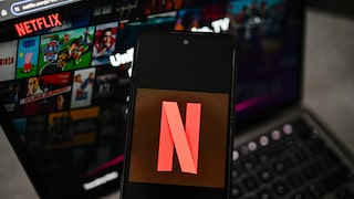 A smartphone displaying the Netflix logo is positioned in front of a laptop. The laptop screen shows the Netflix homepage with various movie and series tiles.