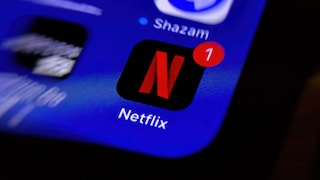 Close-up of a smartphone screen displaying the Netflix app icon. The red Netflix logo on a black background is centered and features a red notification badge with the number 1. Above it is the Shazam app icon on a blue background. The screen is blurred, showing a strong zoom effect.