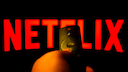A hand holds a remote control in front of a screen displaying a large red Netflix logo on a black background.