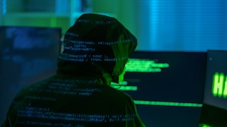 A hooded figure sits in front of several computer screens in a dimly lit room. Blurred lines of code are visible on their clothing and the monitors. The scene is bathed in green and blue light.