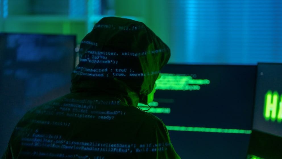 A hooded figure sits in front of several computer screens in a dimly lit room. Blurred lines of code are visible on their clothing and the monitors. The scene is bathed in green and blue light.