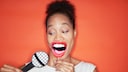 A person stands against an orange background, holding a prop shaped like a large mouth in front of their face, with a stylized cardboard microphone beside it. The image symbolizes speech, synchronization, and the adaptation of lip movements.