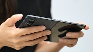 A person is holding an open Samsung Galaxy Z Fold 8 with both hands. The back with the camera module and the hinge of the foldable smartphone are visible. The background is blurred.