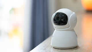 White smart home camera with a black lens cover on a light wood surface. In the background, blurred curtains and warm light sources are visible in an interior setting.