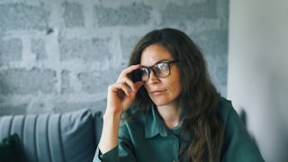 The image shows a person with dark long hair and wearing black glasses. One hand is on the glasses' frame while the person sits at a table. In the background, there is a light gray textured wall and a cushioned bench. The person is wearing a dark green top. The photo is in landscape format, with the person primarily occupying the right half of the image.