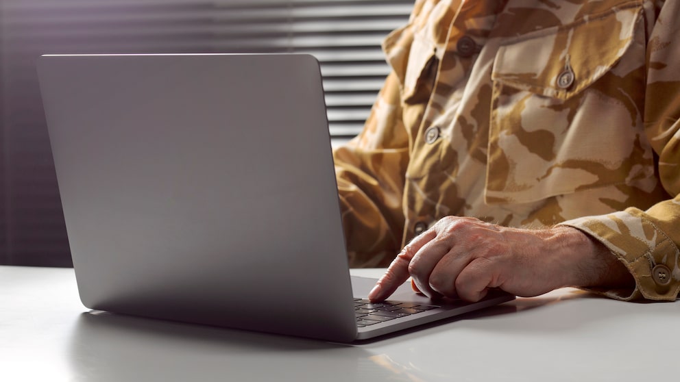 Close-up of an open laptop on a desk. Next to it, hands and the upper body of a person in camouflage uniform are visible, working on the keyboard. In the background, there are horizontal slats of a window or blinds.
