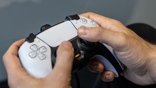 Two hands hold a black-and-white PlayStation controller with buttons and analog sticks against a gray background.