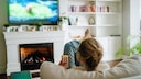 A person sits on a sofa in a living room, looking at a blurred television. They hold a bag of popcorn in their hand. In front of the sofa is a fireplace, with plants and decorations on a shelf beside it.