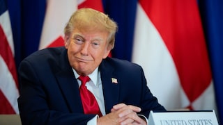 U.S. President Donald Trump sits in a dark suit with a red tie at a table in front of several flags. In front of him is a nameplate that reads "United States."