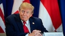U.S. President Donald Trump sits in a dark suit with a red tie at a table in front of several flags. In front of him is a nameplate that reads "United States."