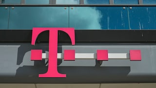 Magenta-colored Telekom logo on the facade of a modern office building. The logo is mounted in front of a glass facade with dark window panes. Clouds are reflected in the glass at the top of the image.