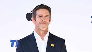 A person in a dark blue blazer and white shirt stands in front of a light-colored event backdrop. Behind their head, a black Apple logo is visible. On the lapel of the blazer is a yellow badge with the word "BELIEVE." Blue lettering is partially visible at the edges of the image.