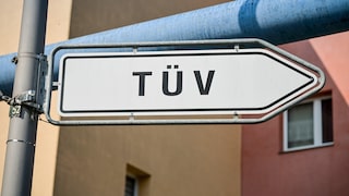 White arrow-shaped sign with the inscription "TÜV" on a metal pole in front of buildings and a blue pipeline in the background