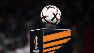 In the foreground, a white soccer ball with black and orange elements is visible. The ball rests on a black pedestal featuring an orange design and the UEFA Europa League logo. The background is dark and blurred, with people only faintly discernible.