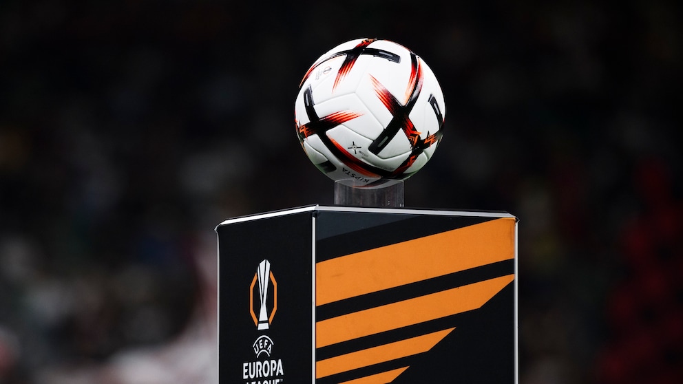 In the foreground, a white soccer ball with black and orange elements is visible. The ball rests on a black pedestal featuring an orange design and the UEFA Europa League logo. The background is dark and blurred, with people only faintly discernible.