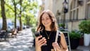 A person with shoulder-length hair stands on a sunny sidewalk between trees and buildings, looking at a smartphone in their hand. A light fabric bag hangs over one shoulder, and in the blurred background, a street, bicycles, and building facades can be seen. Share your feedback on BizChat.