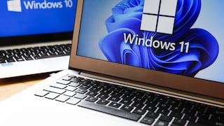 Two open laptops are side by side on a table. The screen in the foreground displays the Windows 11 logo against a blue background. In the background, another screen shows the Windows 10 logo on a blue background. Both devices are slightly offset from each other.