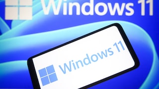 A smartphone displays the Windows 11 logo in front of a screen featuring a large Windows 11 inscription against a blue background.