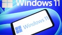 A smartphone displays the Windows 11 logo in front of a screen featuring a large Windows 11 inscription against a blue background.