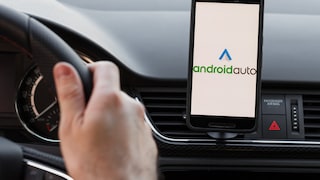 A person is sitting in a car with their hands on the steering wheel. A phone is visible in a dashboard mount, displaying Android Auto.