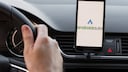 A person is sitting in a car with their hands on the steering wheel. A phone is visible in a dashboard mount, displaying Android Auto.
