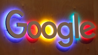 A large Google logo made of white three-dimensional letters is mounted on a wooden wall. Behind each letter, colored light panels in blue, red, yellow, and green illuminate the wall. The photo captures the logo head-on, occupying most of the image.