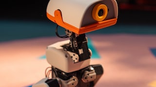 Close-up of the MicroDuck robot featuring a white casing, orange details, and a camera in its head. Mechanical joints and cables are visible.