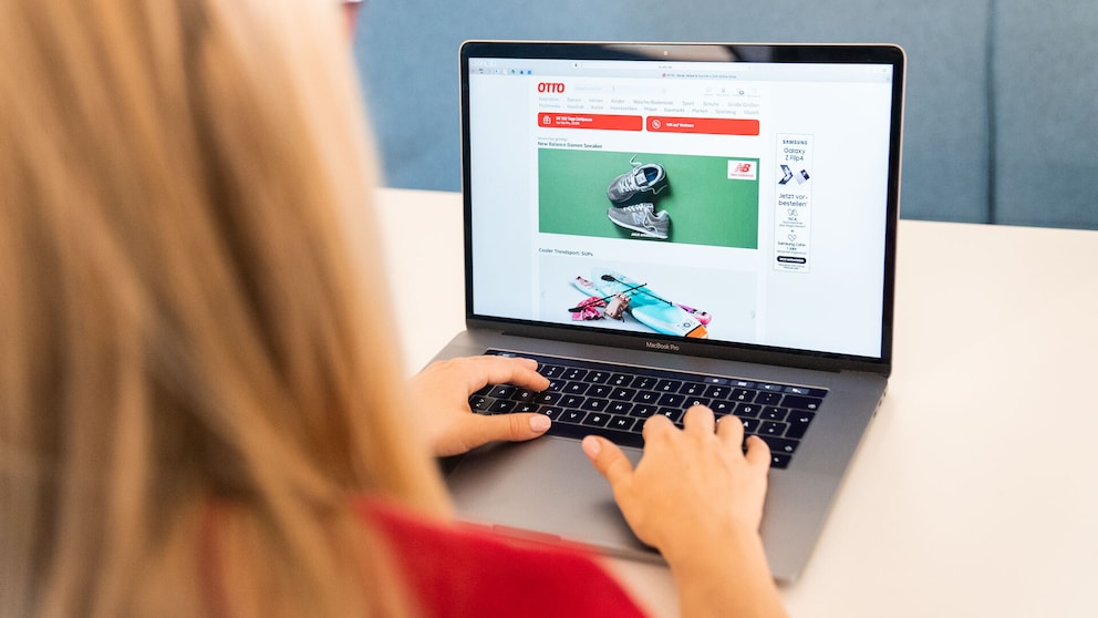 The snippet shows a person sitting at a MacBook Pro with the webpage of the online retailer Otto open. Various products and offers from the Otto online shop are visible on the screen. The person is in the foreground, using the laptop with both hands. In the background, a plain blue wall can be seen.