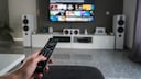 Only a person's hand holding a remote control is visible. It seems the TV on the living room wall was just turned on.