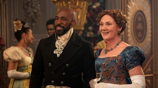 Two people in historical attire stand in a festively decorated hall. One person is wearing a dark jacket with a white cravat, while the other is dressed in a blue gown with copper embellishments, white gloves, and a matching necklace. In the background, more guests in Regency-style clothing can be seen, along with intricately designed wall decorations and floral arrangements. The scene is captured in warm lighting.