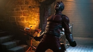 A person in a dark red superhero costume with a mask stands in a brick-walled room under a rounded arch. The figure holds a baton in one hand. The room is lit with warm orange light on the left and cooler light on the right. In the background, a staircase, construction lights, and other technical equipment are visible.