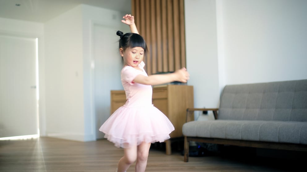 A young girl with dark, tied-up hair dances in a bright living room. She wears a pink ballet dress with a tulle skirt and matching ballet shoes. One arm is raised above her head, while the other is extended to the side. In the background, there is a gray sofa and a wooden dresser.