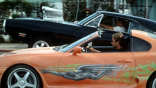 Two sports cars are driving side by side on a road. In the foreground is an orange car with an open roof and striking green and gray side paint. A stylized robot figure is depicted on the driver's door. The driver is visibly seated behind the wheel. Next to it is a black muscle car with a large chrome engine setup on the hood. In the background, buildings, trees, and other street elements can be seen in a blur.
