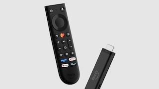 Against a gray background, an Amazon Fire TV Stick and its accompanying remote control are prominently displayed. The black remote features a directional pad, volume buttons, several function keys, and quick access buttons for Prime Video, Netflix, YouTube, and Disney+. Next to it, the black Fire TV Stick with an HDMI connection and the "fire tv" label is visible. Both devices are slightly angled and appear to be floating against the backdrop.