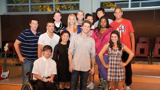 A group photo of 13 cast members from the TV series "Glee" in a room with a small stage. The individuals are standing in several rows next to each other, smiling at the camera. In the foreground, a young man wearing glasses and a white shirt is seated in a wheelchair. Behind the group, chairs, a guitar, and large windows are visible.