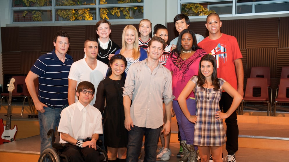 A group photo of 13 cast members from the TV series "Glee" in a room with a small stage. The individuals are standing in several rows next to each other, smiling at the camera. In the foreground, a young man wearing glasses and a white shirt is seated in a wheelchair. Behind the group, chairs, a guitar, and large windows are visible.