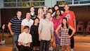 A group photo of 13 cast members from the TV series "Glee" in a room with a small stage. The individuals are standing in several rows next to each other, smiling at the camera. In the foreground, a young man wearing glasses and a white shirt is seated in a wheelchair. Behind the group, chairs, a guitar, and large windows are visible.