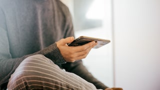 A person sits in a bright room, holding a smartphone horizontally in one hand. The person is wearing a dark gray top and gray striped pants. Their gaze is fixed on the smartphone, with a bright and blurred background.