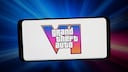 A smartphone is positioned horizontally against a blue and pink glowing background. On the white display, the logo for "Grand Theft Auto VI" is visible. Behind the text, a large Roman numeral six features a gradient transitioning from blue to pink to orange.