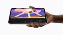 A hand holds an iPad mini in landscape orientation against a white background. The display shows animation software with a timeline and several editing tools open. Above it is a colorful drawing featuring pink, yellow, and violet brushstrokes. The tablet has a black display frame and a light metal casing.