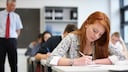 A red-haired student sits at a desk in the classroom, writing with a pen on paper. In the background, other students are seated at their desks, while a teacher wearing a red tie stands in the room.