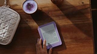 A hand touches a purple Amazon Kindle e-reader on a dark wooden table. The display shows a page of German text. Next to the e-reader are a cup with a light purple drink and foam, and a light-colored woven handbag. Sunlight and shadow patterns fall across the table. The fingernails of the hand are painted purple to match the e-reader.