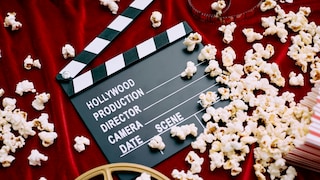 A clapperboard labeled "Hollywood Production" rests on a red fabric background. Scattered around the clapperboard are numerous pieces of popcorn. At the bottom of the image, part of a film reel is visible, while at the top, there's a strip of film. To the right, there's a red-and-white striped popcorn container. The scene is photographed from above, showcasing various cinema and film props.
