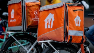 Several orange Lieferando delivery bags with white logos and gray reflector stripes are mounted on the luggage racks of bicycles. One of the bags also features the UEFA Champions League logo. In the blurred background, there are several people, including one person in a yellow jacket.
